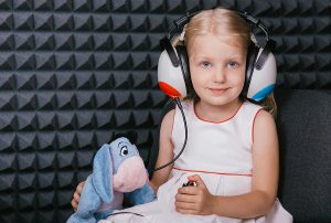 Smiling little girl holding a stuffed animal and wearing headphones while working with a pediatric audiologist in a soundproof room