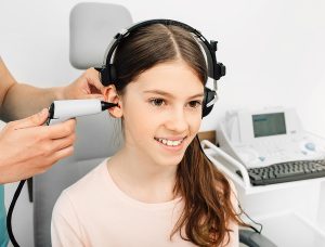 Smiling young girl wearing headphones while a pediatric audiologist tests her hearing